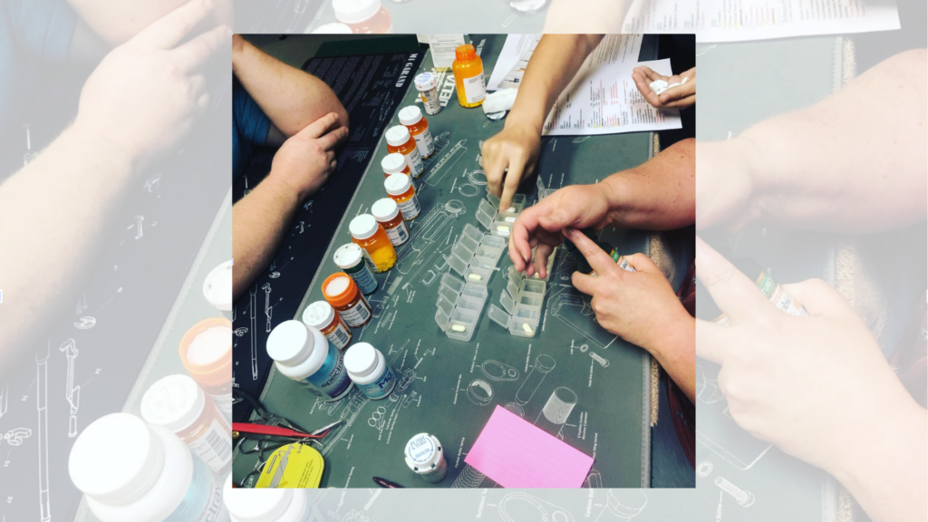 Two people sorting medications into a weekly pill organizer surrounded by multiple prescription bottles and discharge paperwork on a table — a community health worker helping a patient with medication reconciliation after hospital discharge.